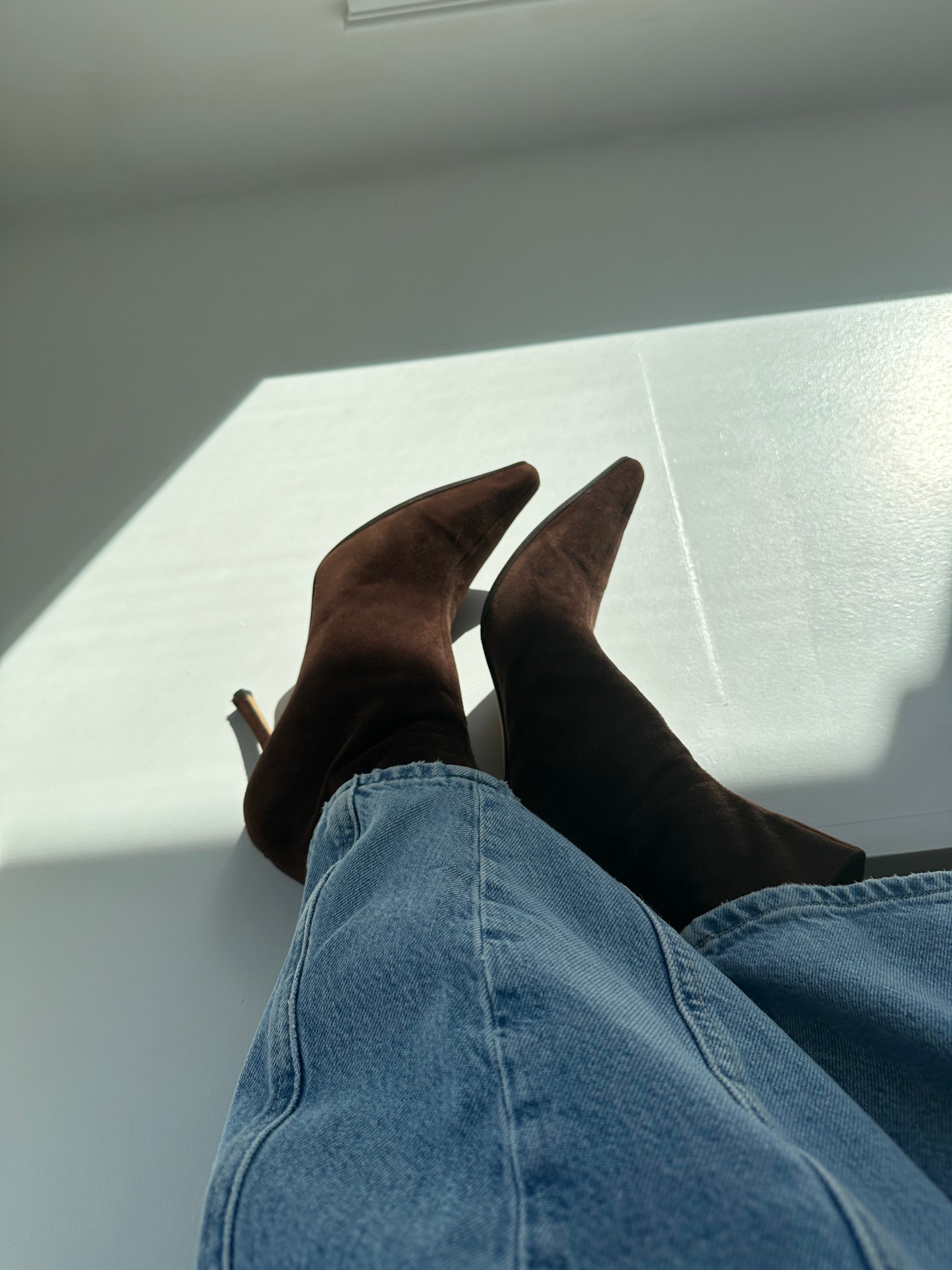 A pair of brown Jimmy Choo booties with pointed toes and a heel, peeping out from rolled-up blue jeans.