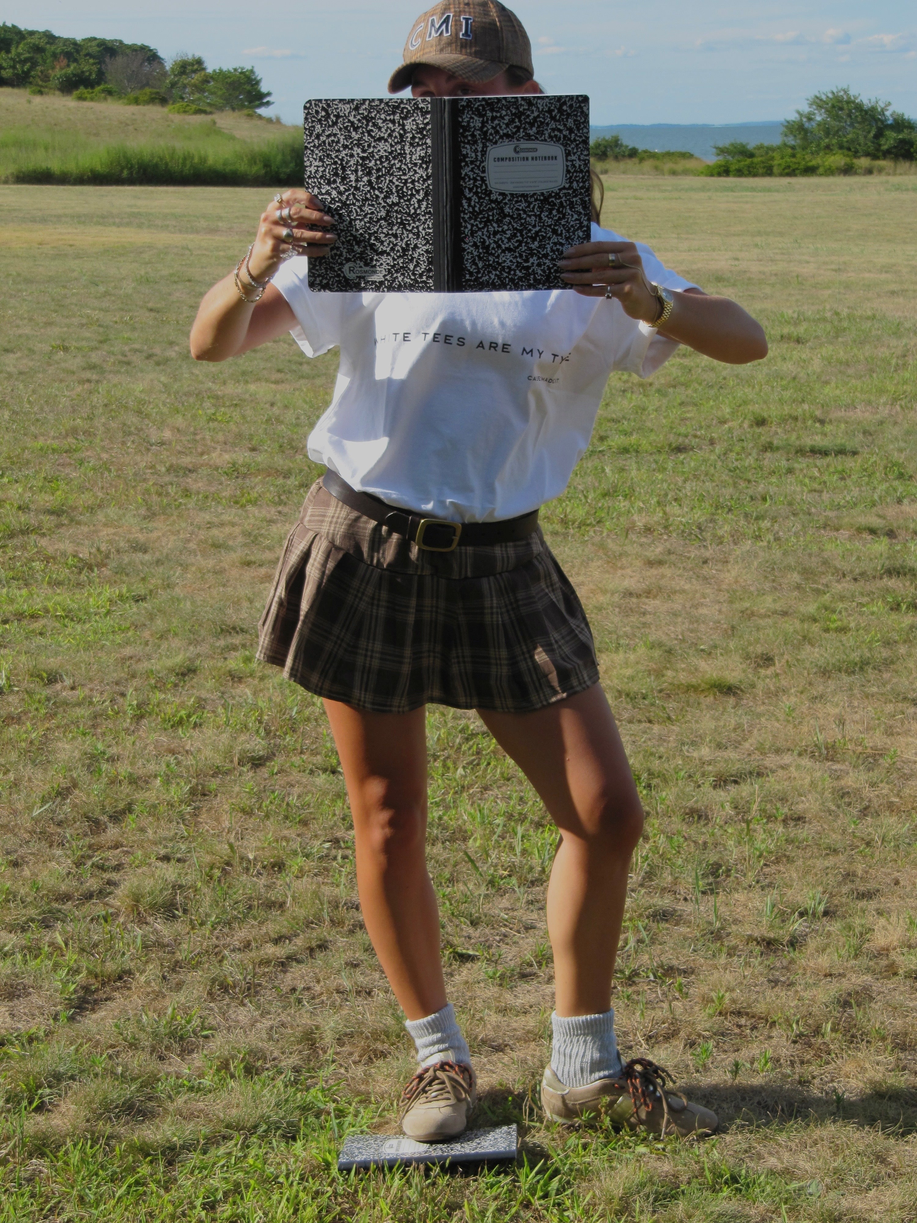 Person in a field holding a book over their face wearing a white t-shirt